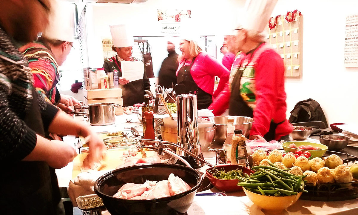 A group of people wearing Christmas sweaters cooking a meal together around a counter. One person is cutting and prepping chicken on a cutting board.